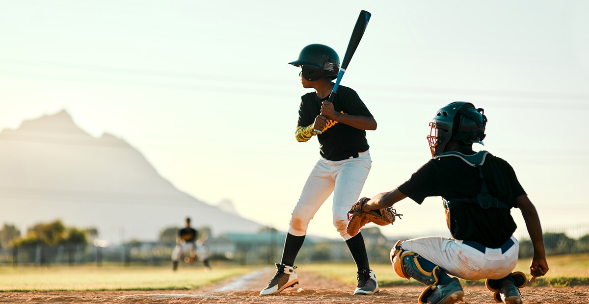 A young batter waits on the pitch, catcher crouched behind the plate